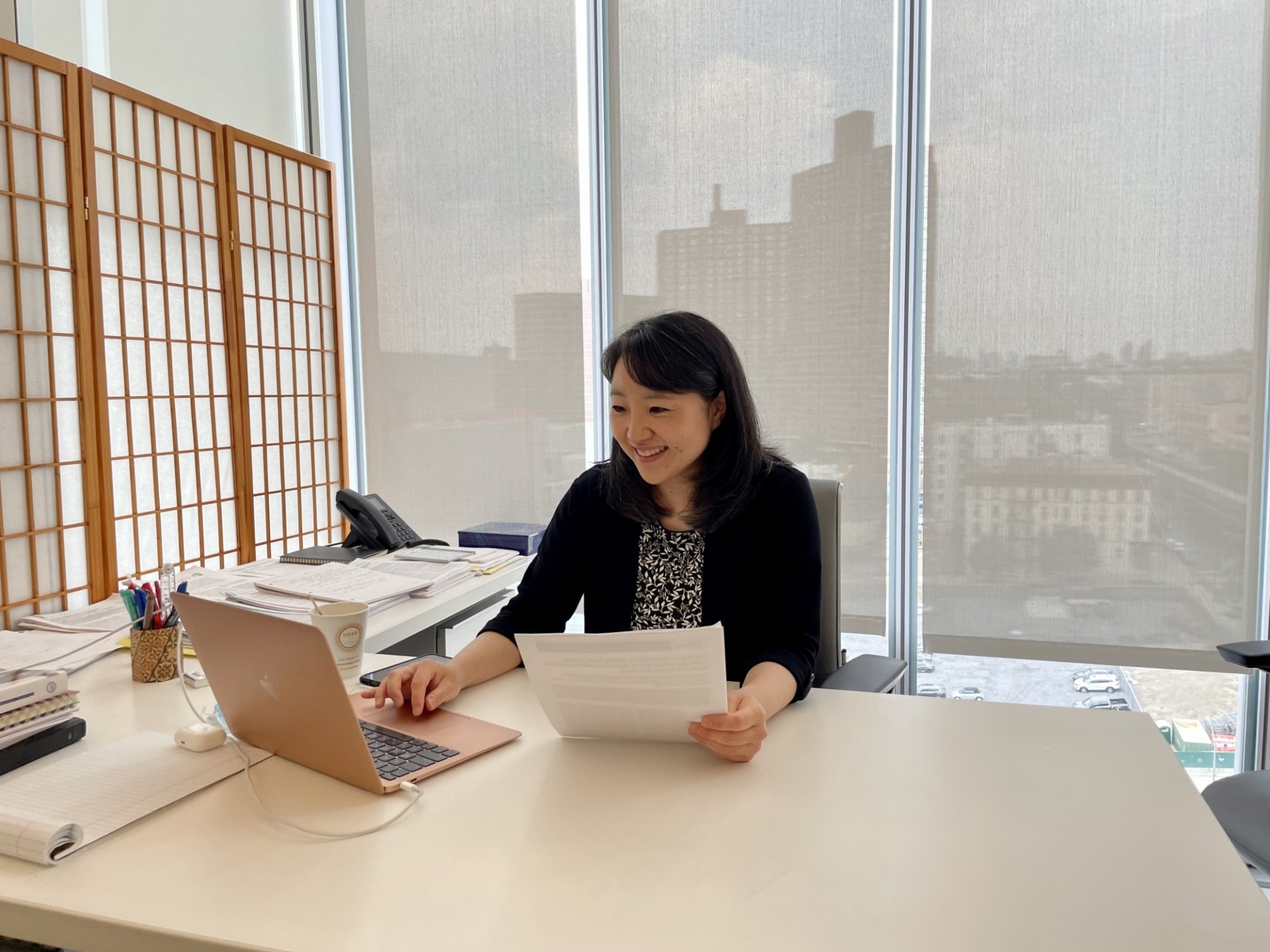 Minoree at her desk, holding a paper and looking at her laptop. 